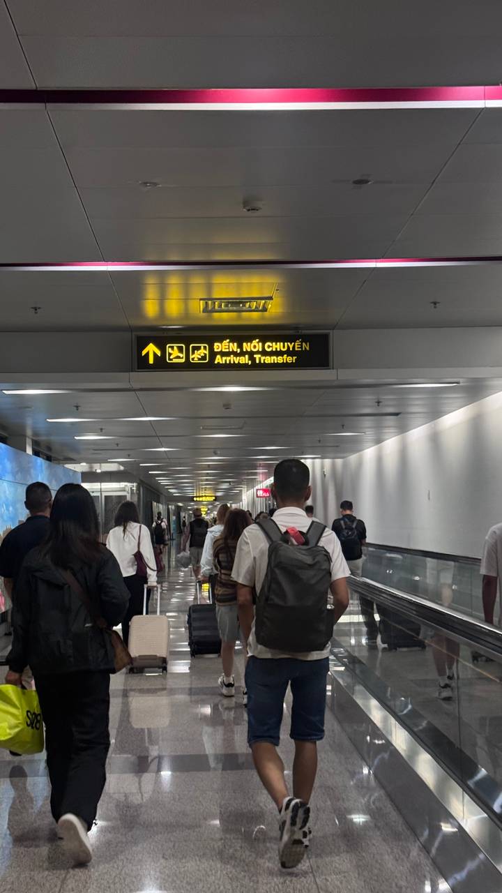 Travelers walk through an airport corridor beneath a yellow bilingual sign for arrivals and transfers.