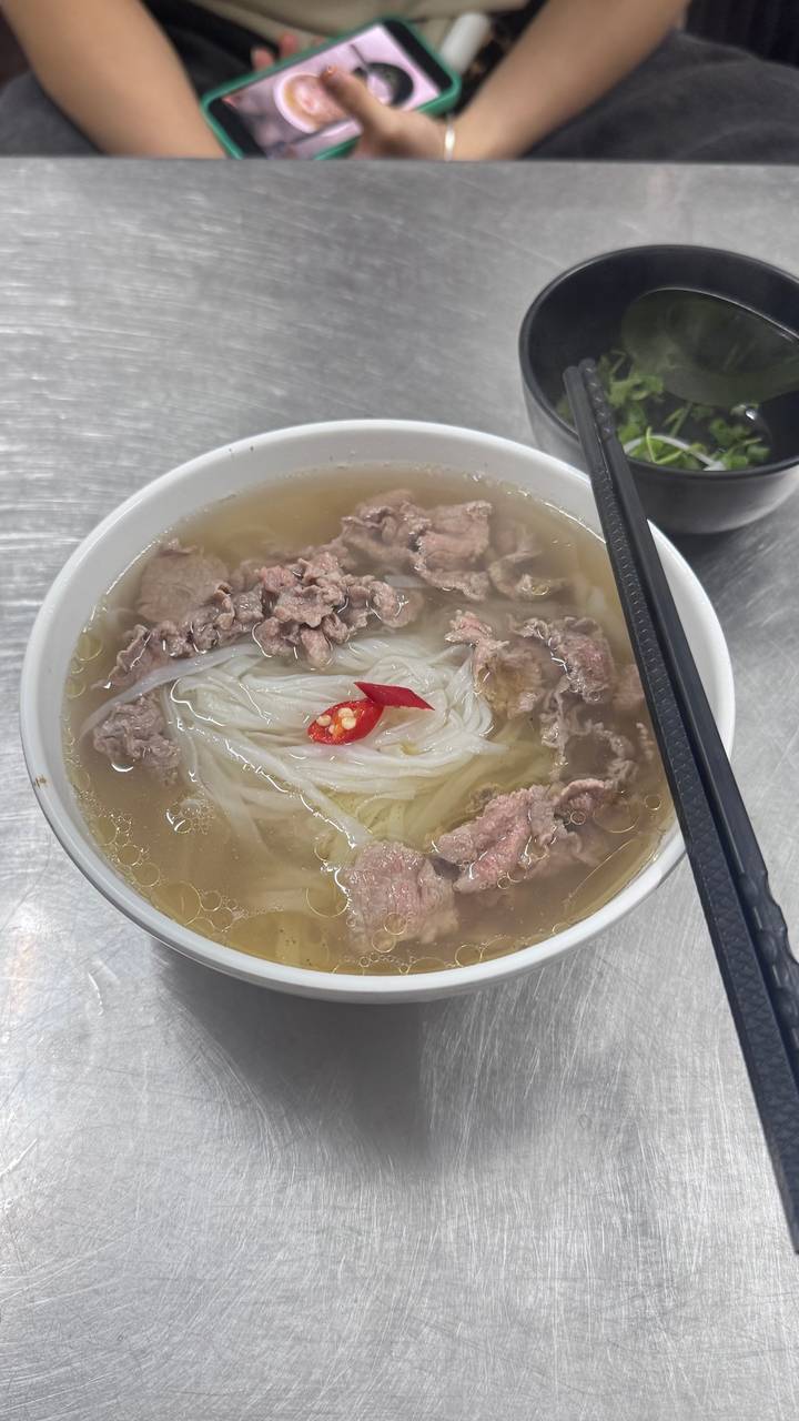 A steaming bowl of Vietnamese pho with beef slices and a red chili garnish, chopsticks resting on the rim.