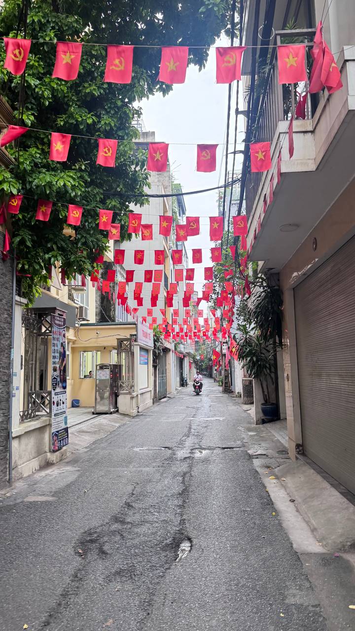 A narrow Hanoi street lined with buildings is decorated with rows of red Vietnamese flags; a motorcyclist rides away in the distance.