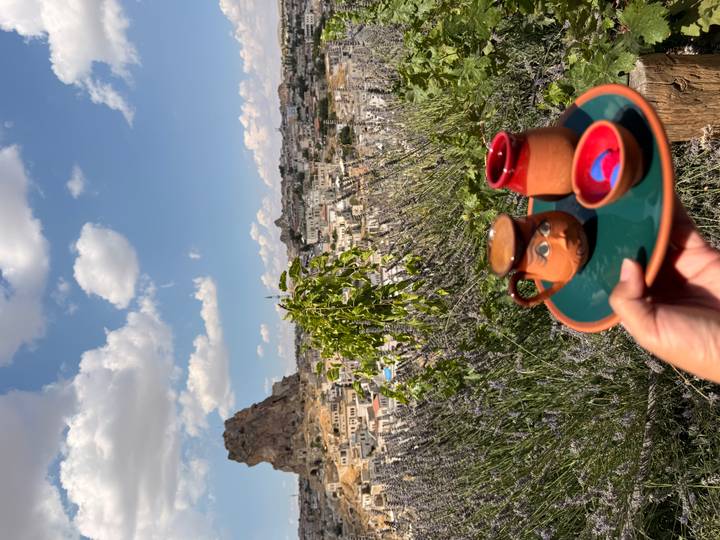 Hand-held clay mugs on a tray in the foreground frame a panoramic view of a Cappadocian town and rocky hill.