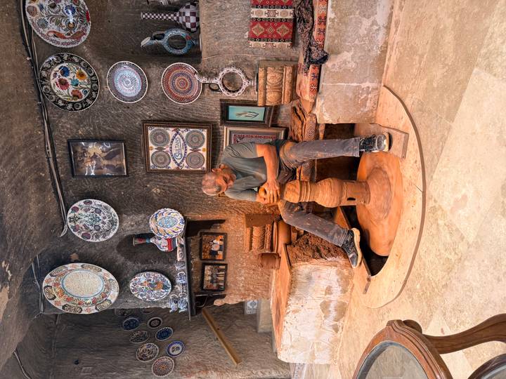 A potter concentrates while shaping clay on a kick wheel inside a workshop adorned with colorful plates.