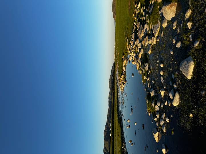 Serene shallow pool with scattered rocks on a grassy plain, distant low mountains under a clear blue sky during warm light.