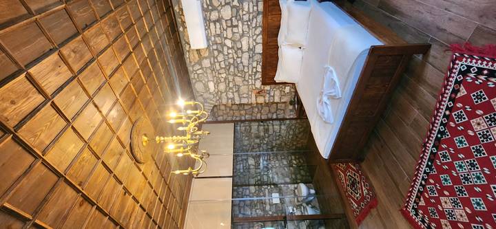 Rustic boutique bedroom with stone walls, wooden ceiling, chandelier and glass-enclosed bathroom.