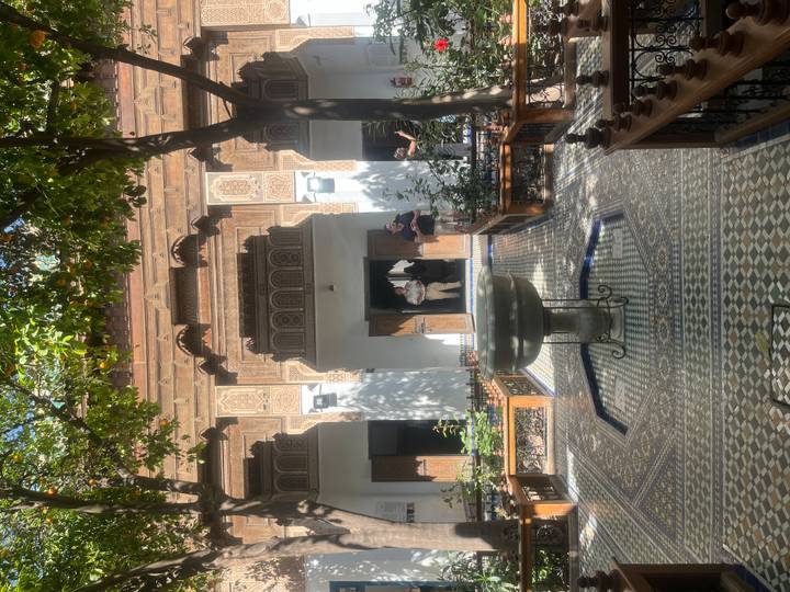 Leafy courtyard of a traditional Moroccan house with tiled fountain and visitors exploring.