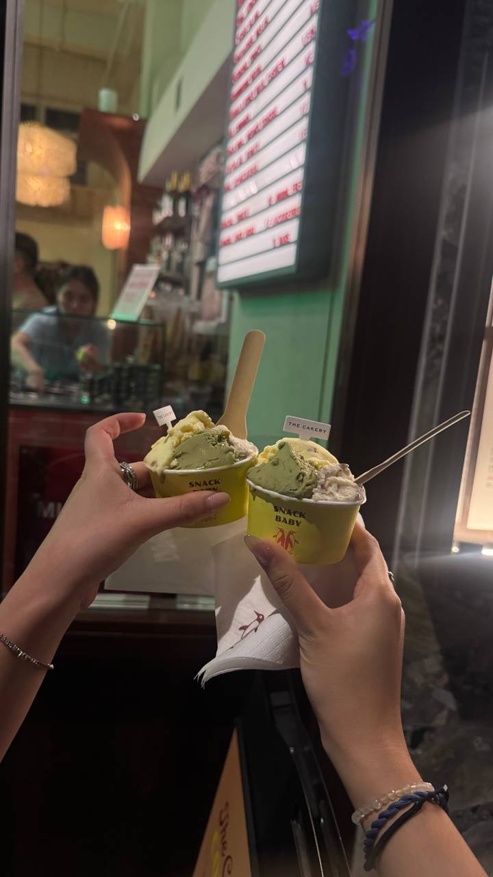 Two hands raise cups of artisanal ice cream inside a softly lit shop at night.