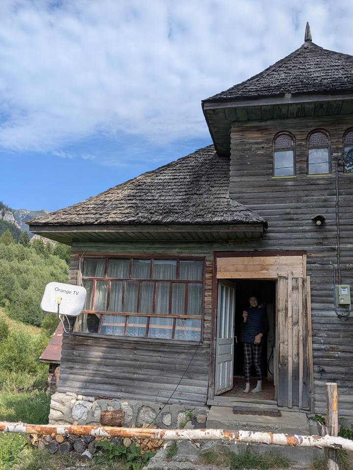 Weathered wooden farmhouse with mismatched windows; a person peeks out from the doorway.