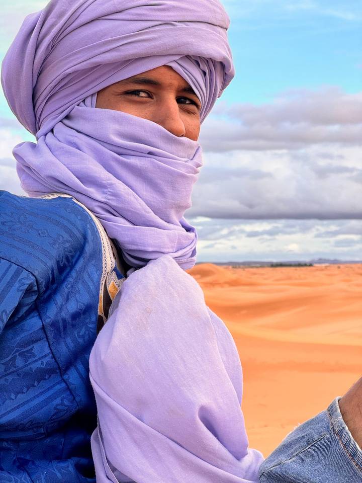 Berber man in blue robe and lavender turban looking across vast orange dunes.