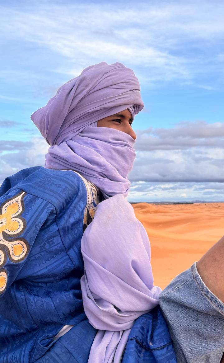 Side profile of turban-clad traveller smiling against sweeping Sahara sandscape.