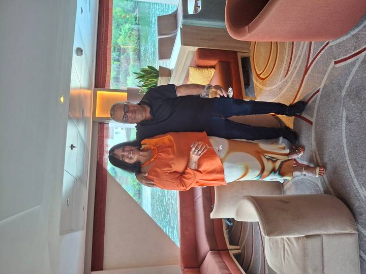 Smiling couple holding wine glasses in a river-cruise lounge with water visible through windows.