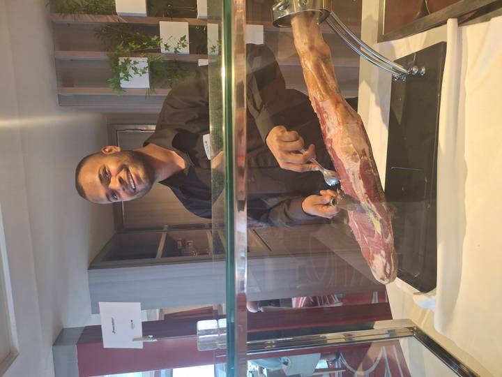 Chef slices cured ham behind a glass counter, smiling at the camera.