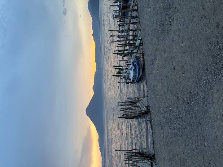 Tranquil lake scene at sunset with a wooden dock, small boat and a volcanic mountain silhouette in the background.