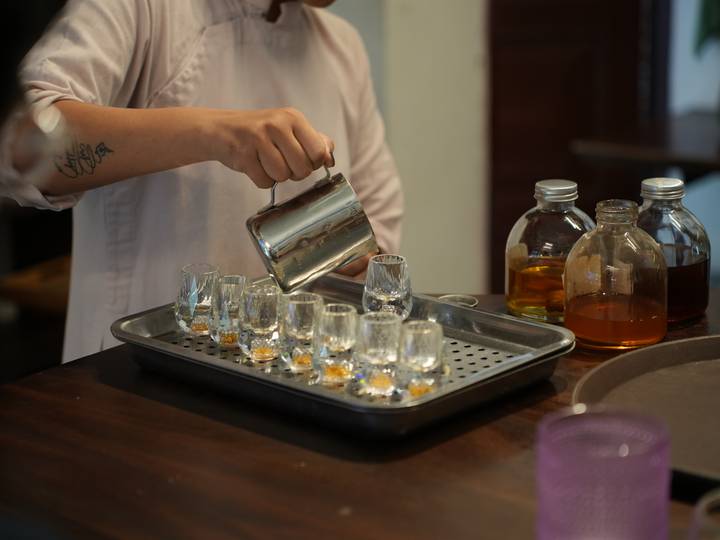 Hand holding a metal pitcher pouring amber coffee into miniature glasses beside syrup bottles.