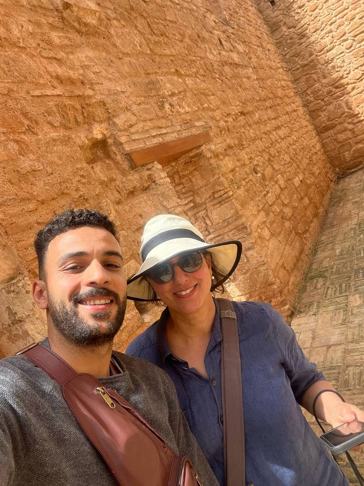 Smiling travellers take a sunny selfie in front of ancient sandstone walls with ornate carvings.
