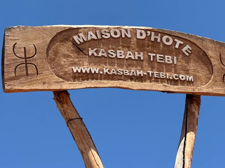 Wooden entrance sign reading 'Maison d'Hôte Kasbah Tebi' set against a bright blue desert sky.