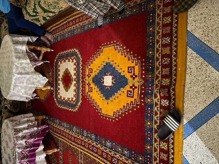 Colorful Berber carpet fills the frame of an indoor room, with patterned tables and a sandal in one corner.