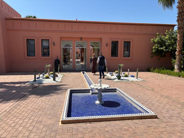 Entrance to a modern riad with tiled fountain and desert plants, two visitors walking toward glass doors.