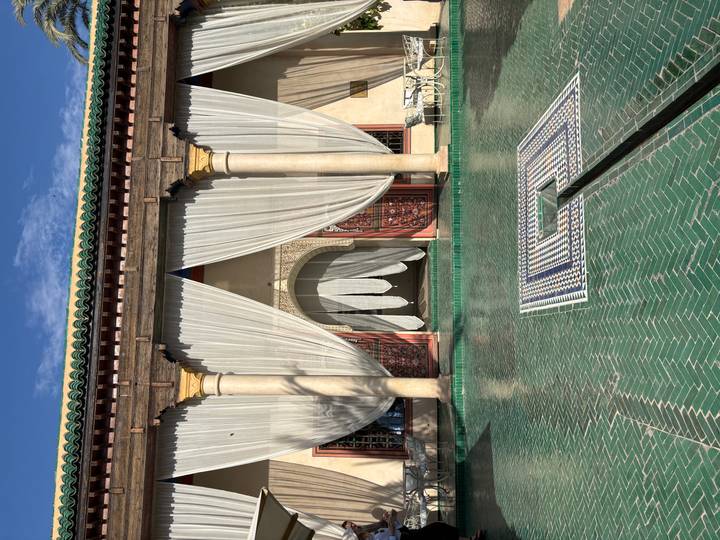 Elegant riad courtyard with green-tiled pool, cream drapes and carved columns highlighted by afternoon sun.