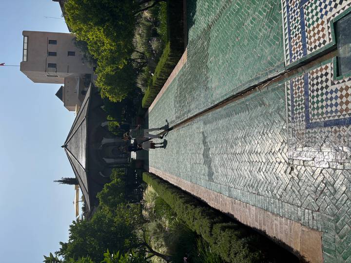 Long tiled pathway bordered by lush gardens leading to a pavilion where two visitors stroll in the sun.
