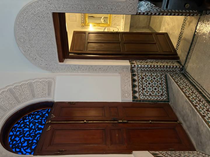 Decorative doorway inside a riad featuring carved plaster, zellige tiles and warm lighting leading to a bathroom.