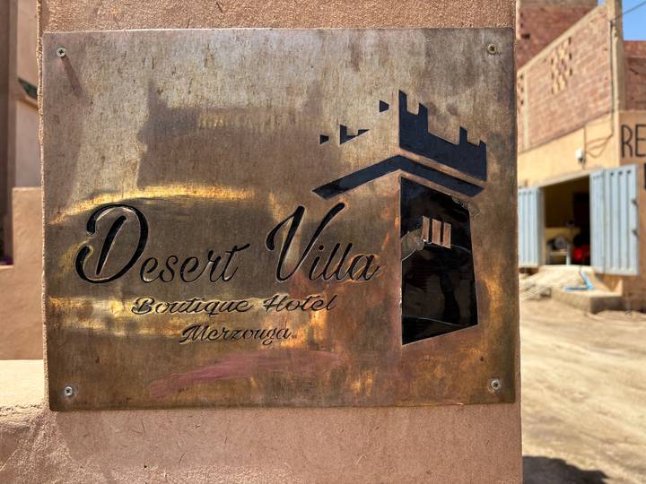 Weathered brass sign for 'Desert Villa Boutique Hotel Merzouga' mounted on a wall in desert light.