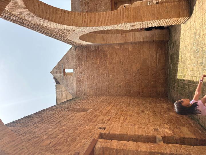 Sunlit ruins with high brick walls and arched openings; a visitor rests on the tiled floor while another explores.