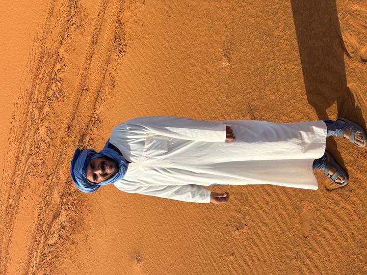 Berber guide in white djellaba and blue turban standing on golden sand with dune tracks behind.