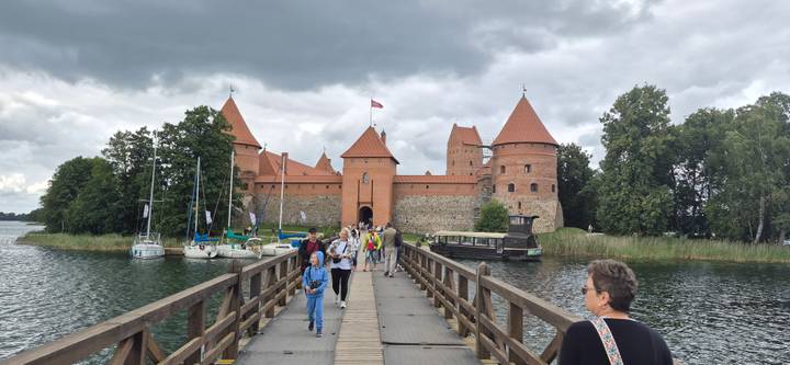 Visitors walk a wooden bridge toward the red-brick towers of Trakai Island Castle surrounded by water and yachts under a cloudy sky.