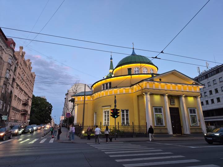 Yellow Orthodox chapel glowing with evening lights on a Riga street at dusk with pedestrians and passing traffic.