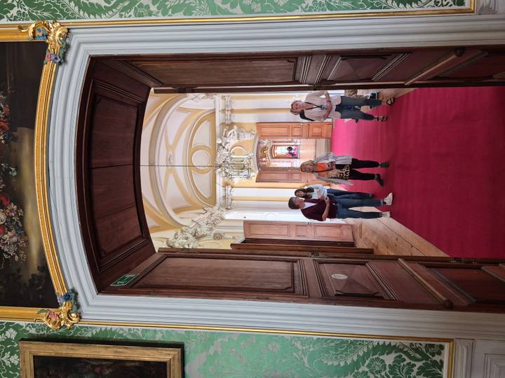Visitors walking through an ornate palace corridor with arched ceilings and a rich red carpet.