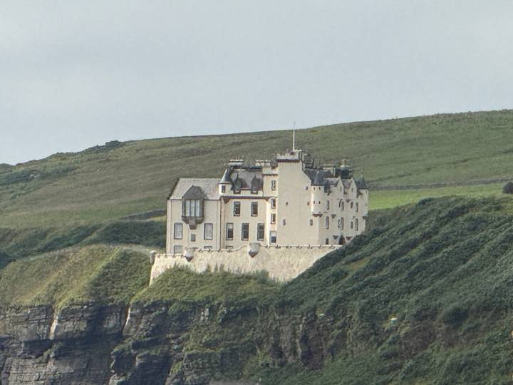 Historic coastal castle perched on a grassy cliff with rolling green hills in the background under a grey sky.