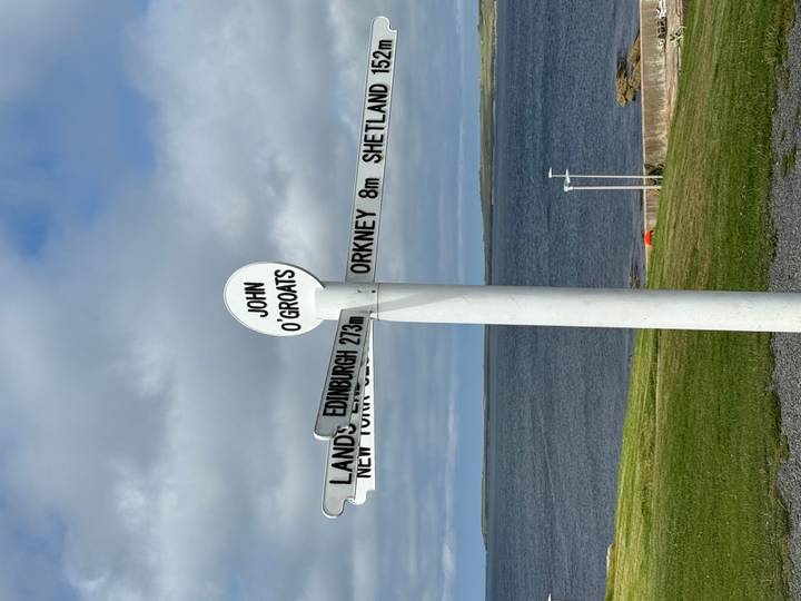 Iconic white signpost at John O'Groats pointing to global destinations with the sea and cloudy sky behind.