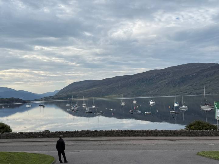 Still lake reflecting moored sailboats with surrounding mountains under a softly overcast sky.
