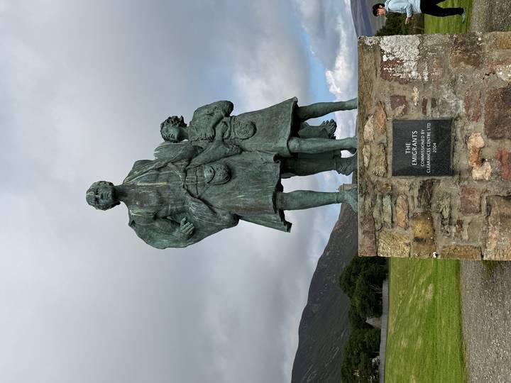 Bronze statue of two Highland emigrants atop a stone plinth with hills and grey clouds in the background.