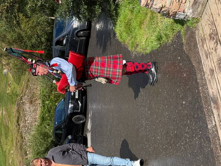 Bagpiper dressed in bright traditional tartan attire playing beside a rural road with parked cars nearby.