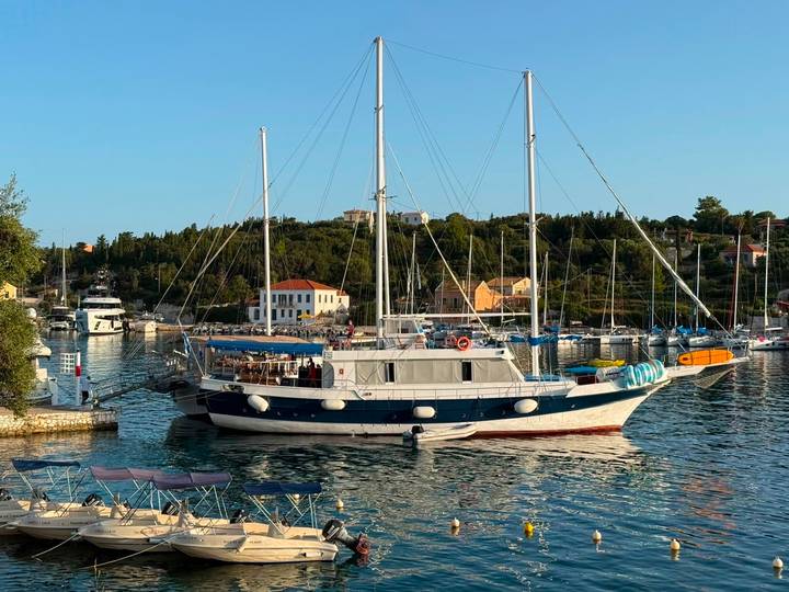 Elegant two-masted yacht moored in picturesque harbor at golden hour with pastel houses and tree-lined hills.