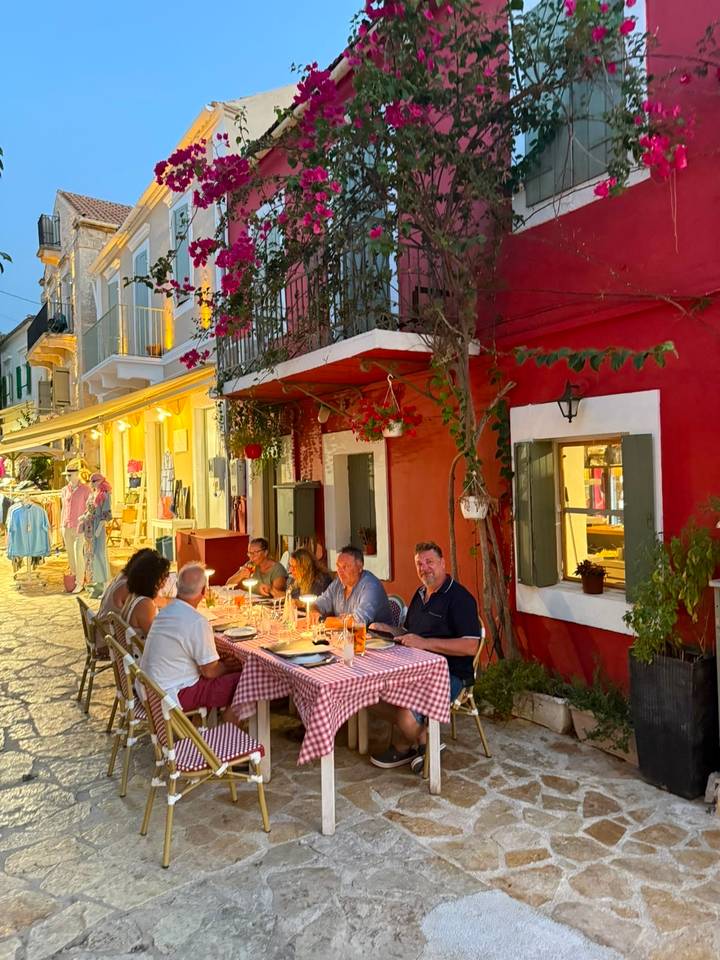 Group dining al fresco at long table outside colorful Mediterranean building lit warmly at dusk.