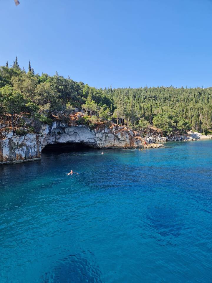 Swimmer enjoying vivid turquoise water near dramatic limestone cave and forested cliffs.