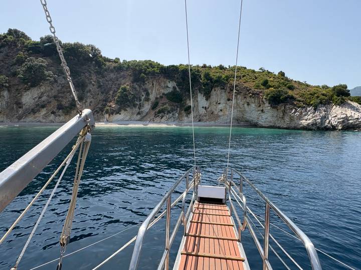 View from sailboat bow toward secluded rocky cove with clear water and green hillside.