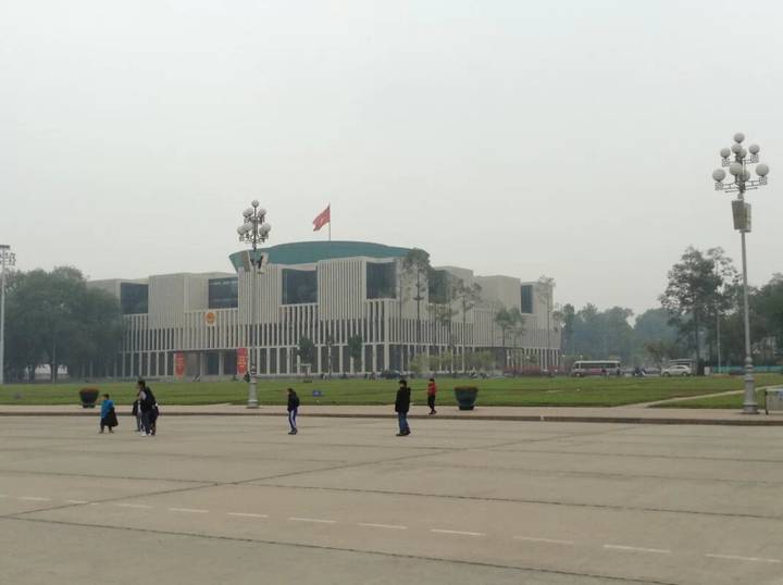 Vietnamese National Assembly building across broad square with flag flying on overcast day, few pedestrians.