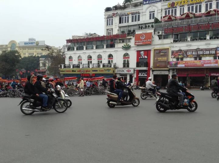 Bustling Hanoi intersection filled with motorbikes, scooters and colonial buildings with signage.