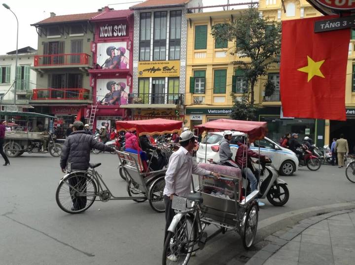 Cyclo drivers with red canopies waiting under Vietnamese flag amid busy street scene.