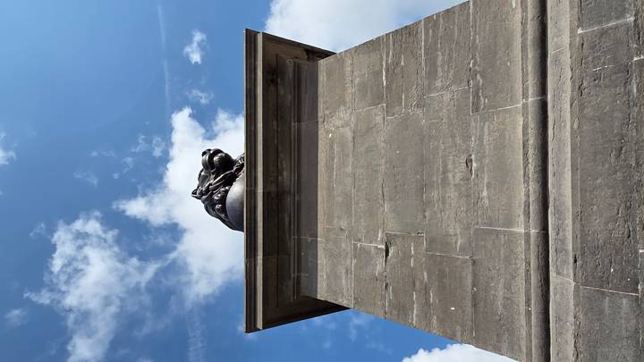 Low-angle view of a stone pedestal topped by a lion statue against a bright blue sky with clouds.