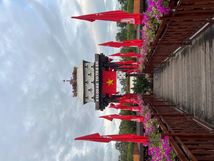Wooden bridge lined with red flags leads to a white gate tower reading 'Welcome to Tra Su'.