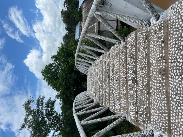 Pebble-patterned footbridge with white railings spans over lush tropical garden under a bright sky.