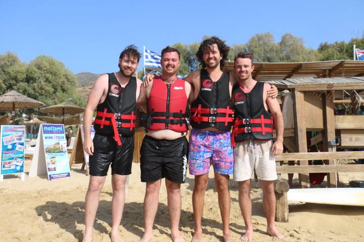 Four men in life jackets stand on a sandy beach after water sports, Greek flag in background.