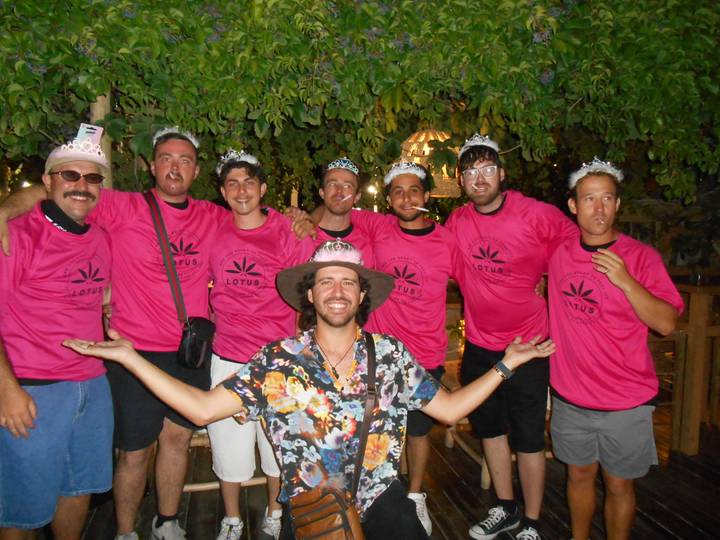 Nighttime group shot of friends wearing matching pink shirts and novelty crowns under leafy trees.