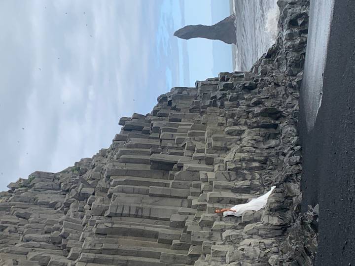 A woman in a white dress stands against dramatic basalt columns on a black-sand beach with the ocean in the background.