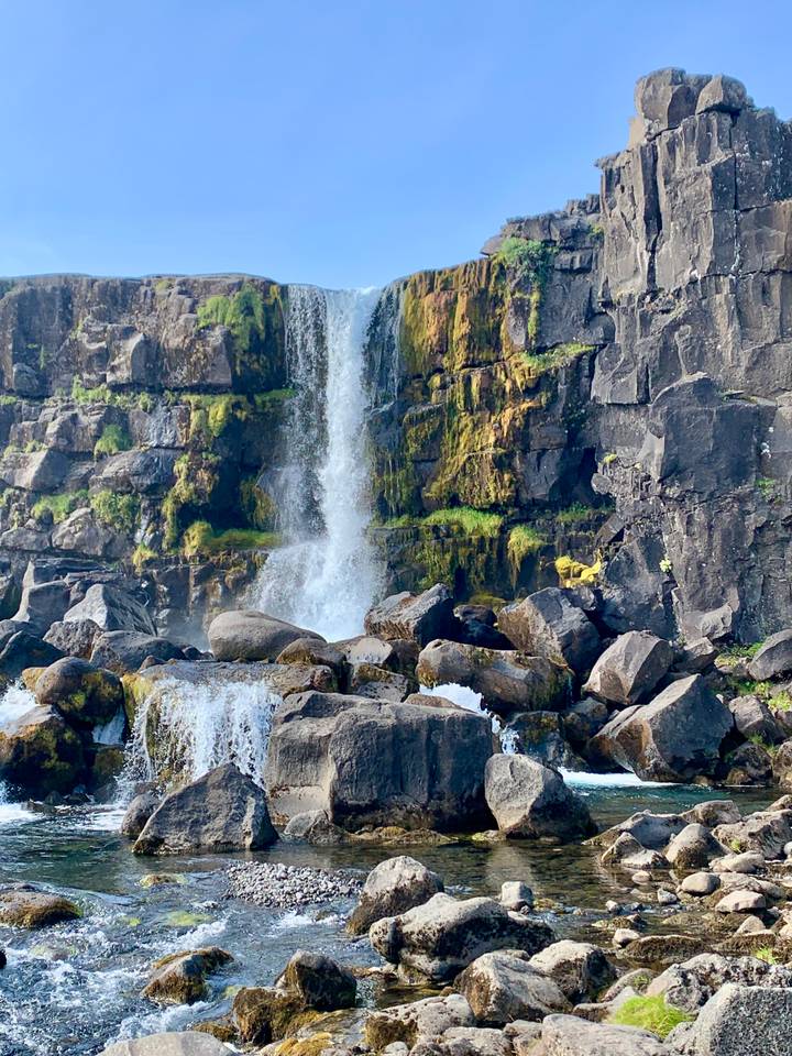 A cascade tumbles over dark volcanic rocks covered in green moss, spraying white water below.