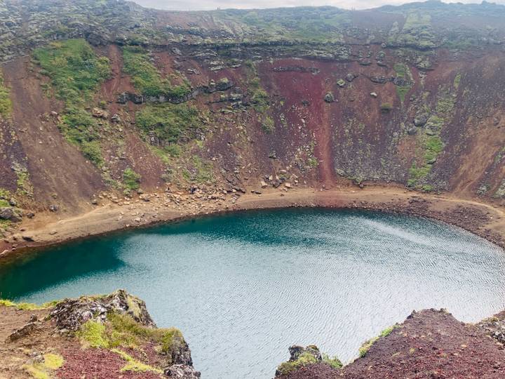 A vivid blue crater lake surrounded by multicolored volcanic rock walls and sparse vegetation.
