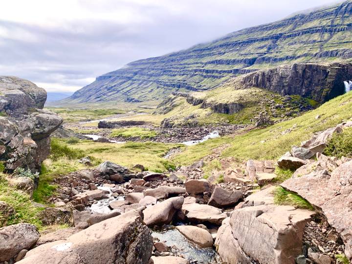 Broad valley with terraced green cliffs, scattered rocks and a small stream winding through the landscape.